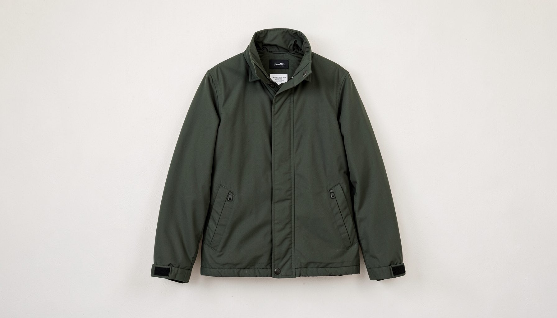 A dark green technical outdoor jacket hanging against a plain off-white wall