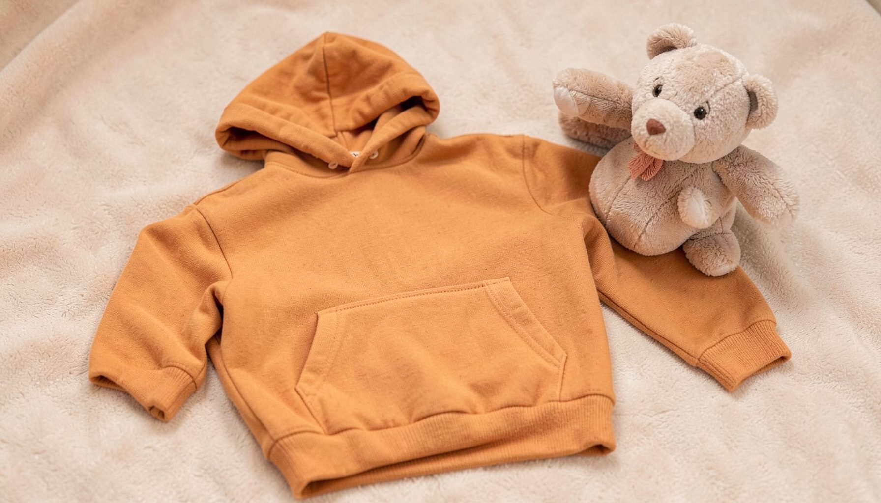 A small child-size hoodie in amber color laid flat next to a soft toy, warm and playful mood